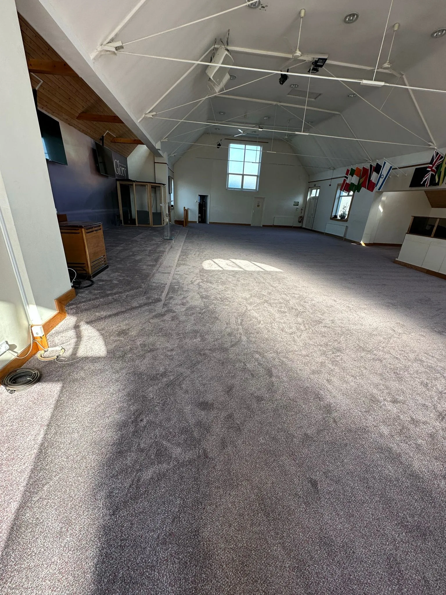 Dark grey carpet installation completed in a church hall with raised stage platform and international flags on wall